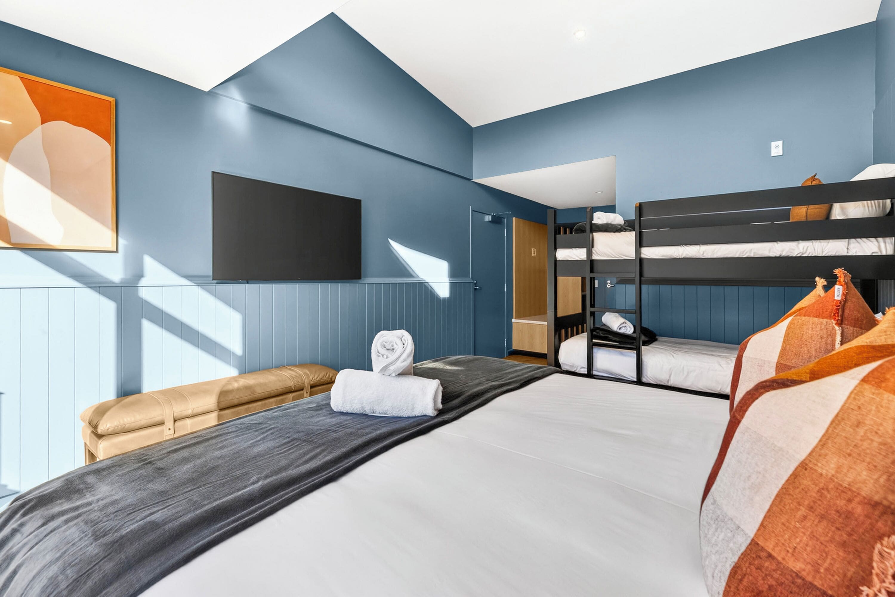 A modern hotel room with a large bed, a sofa, a TV mounted on the wall, and a bunk bed.