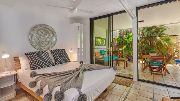 A bedroom with a bed, bedside table, and a view of an outdoor patio with a table and chairs. - Julians Apartments (Byron Bay)