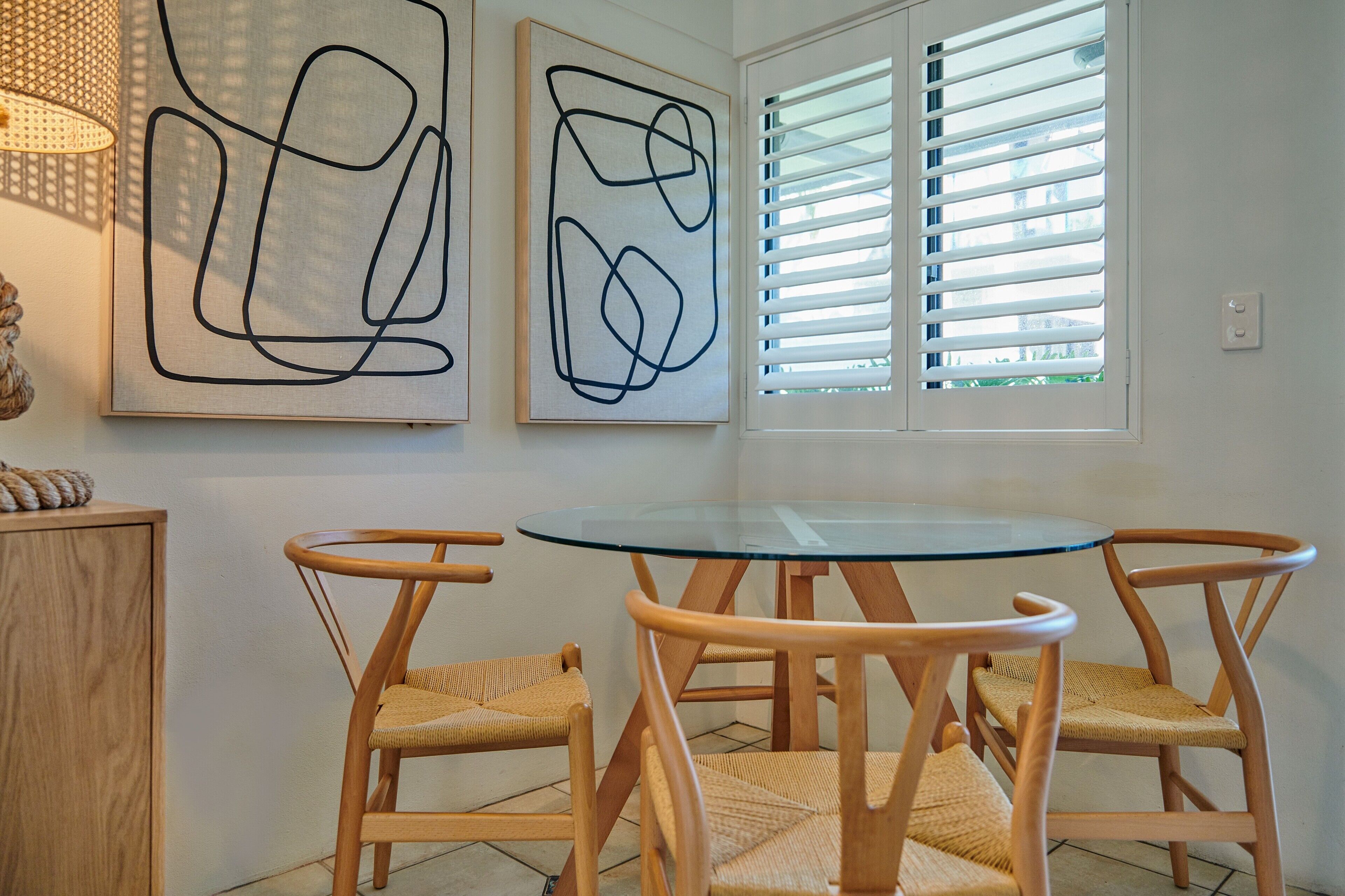 Uma área de jantar com uma mesa redonda de vidro e quatro cadeiras de vime. Há quadros abstratos na parede e uma janela com persianas.