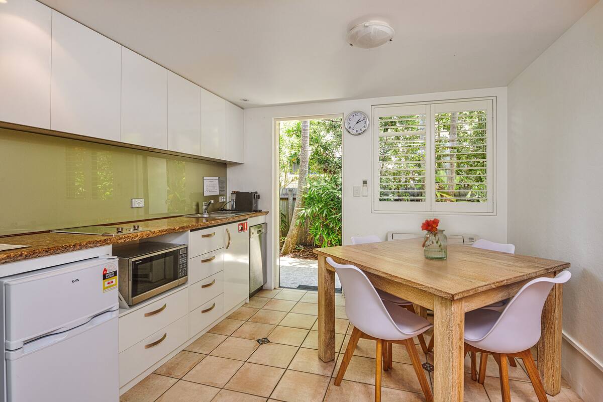 A modern kitchen with white cabinets, a microwave, and a refrigerator. There is a wooden dining table with chairs and a clock on the wall.