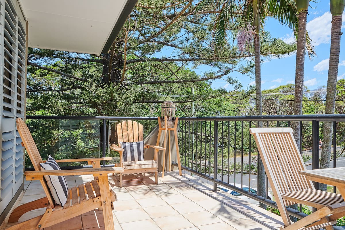 A balcony with wooden chairs and a table, surrounded by a metal railing, overlooking a lush green landscape.