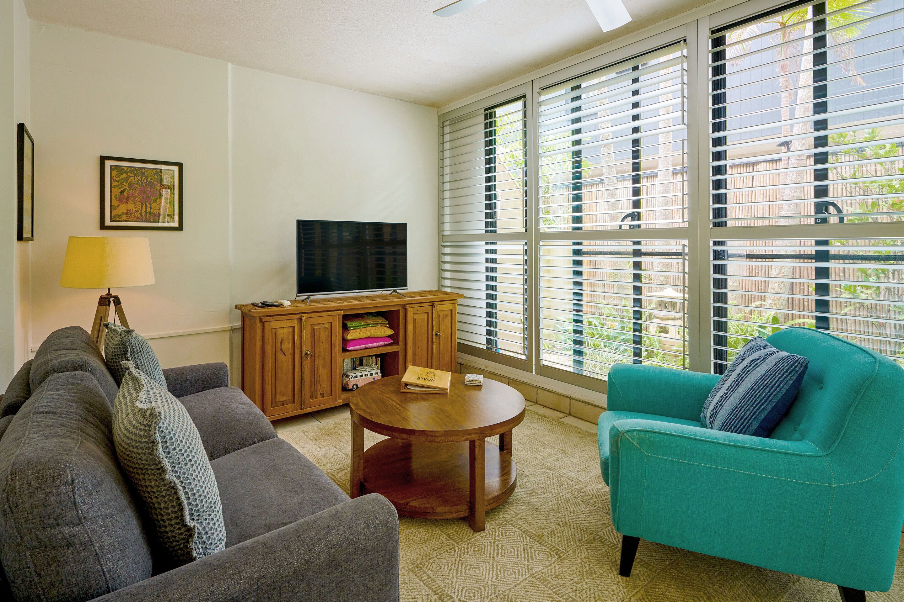 A living room with a sofa, armchair, TV, wooden cabinet, and large windows.