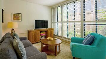 A living room with a sofa, armchair, TV, wooden cabinet, and large windows.