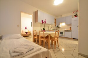 A modern hotel room with a bed, a dining table with chairs, a kitchen area with cabinets and appliances, and a small potted plant.