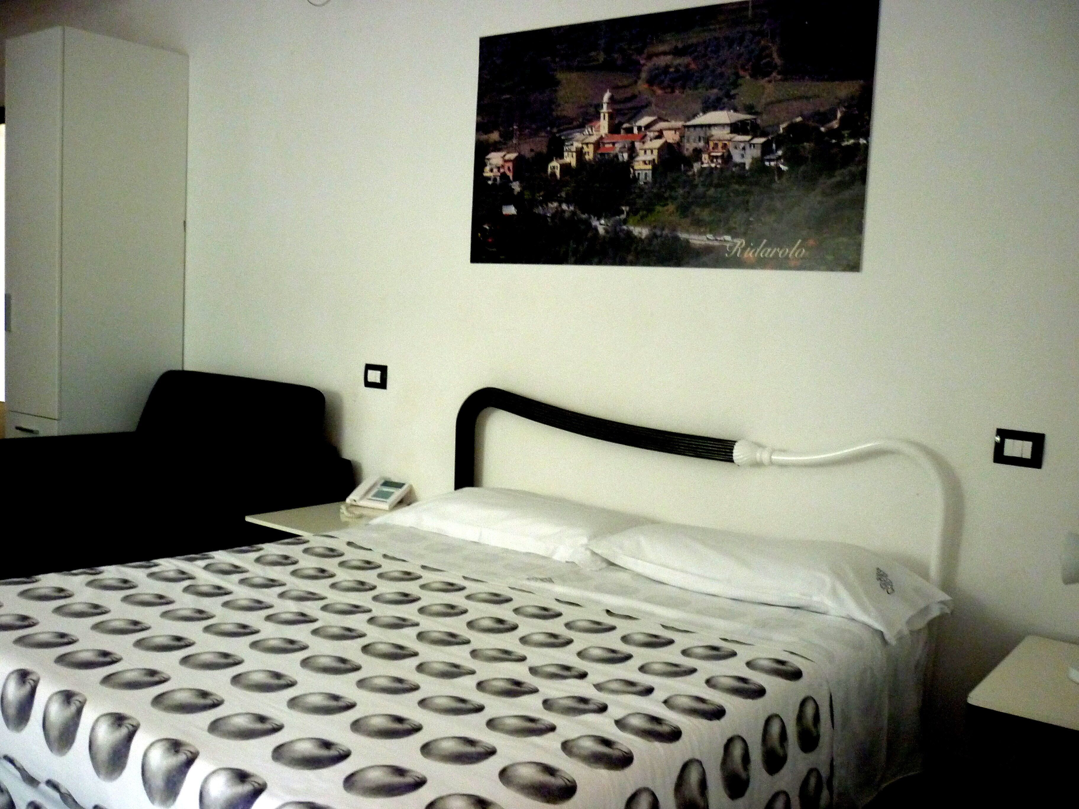 A hotel room with a bed, a nightstand, a telephone, and a painting of a town on the wall.