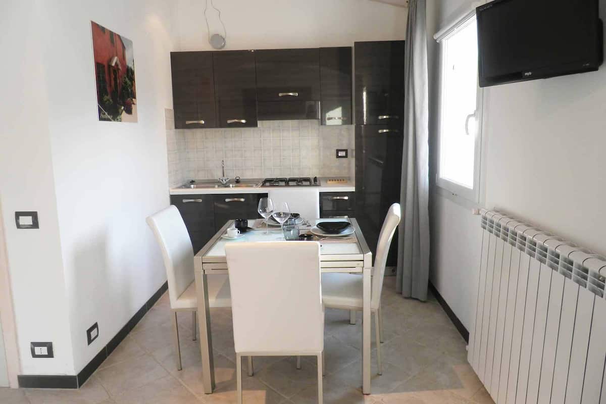 A modern kitchen with dark cabinets, a dining table with chairs, a flat-screen TV mounted on the wall, and a window with curtains.