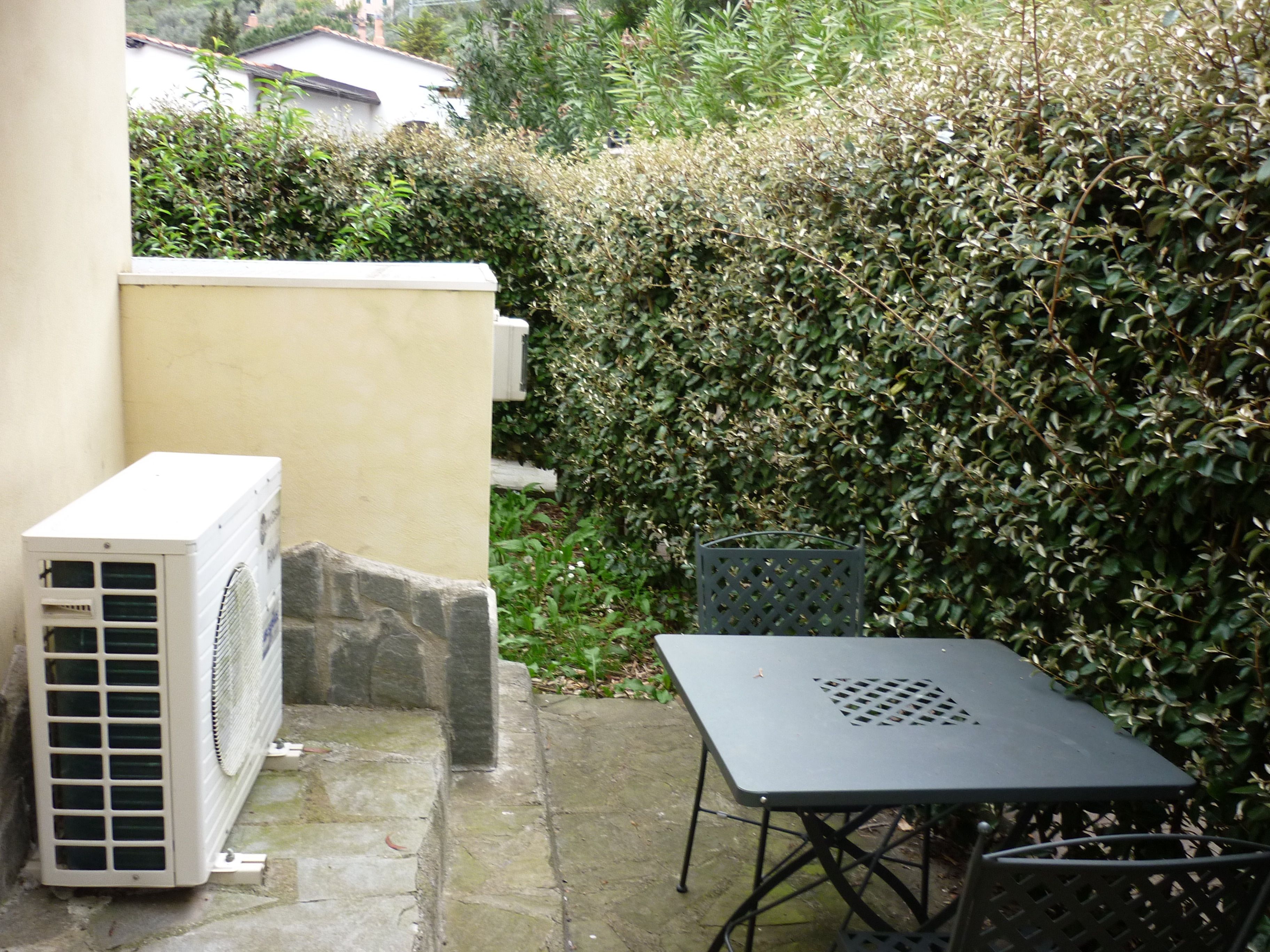 Une terrasse extérieure avec une table et des chaises, un climatiseur blanc et une haie.