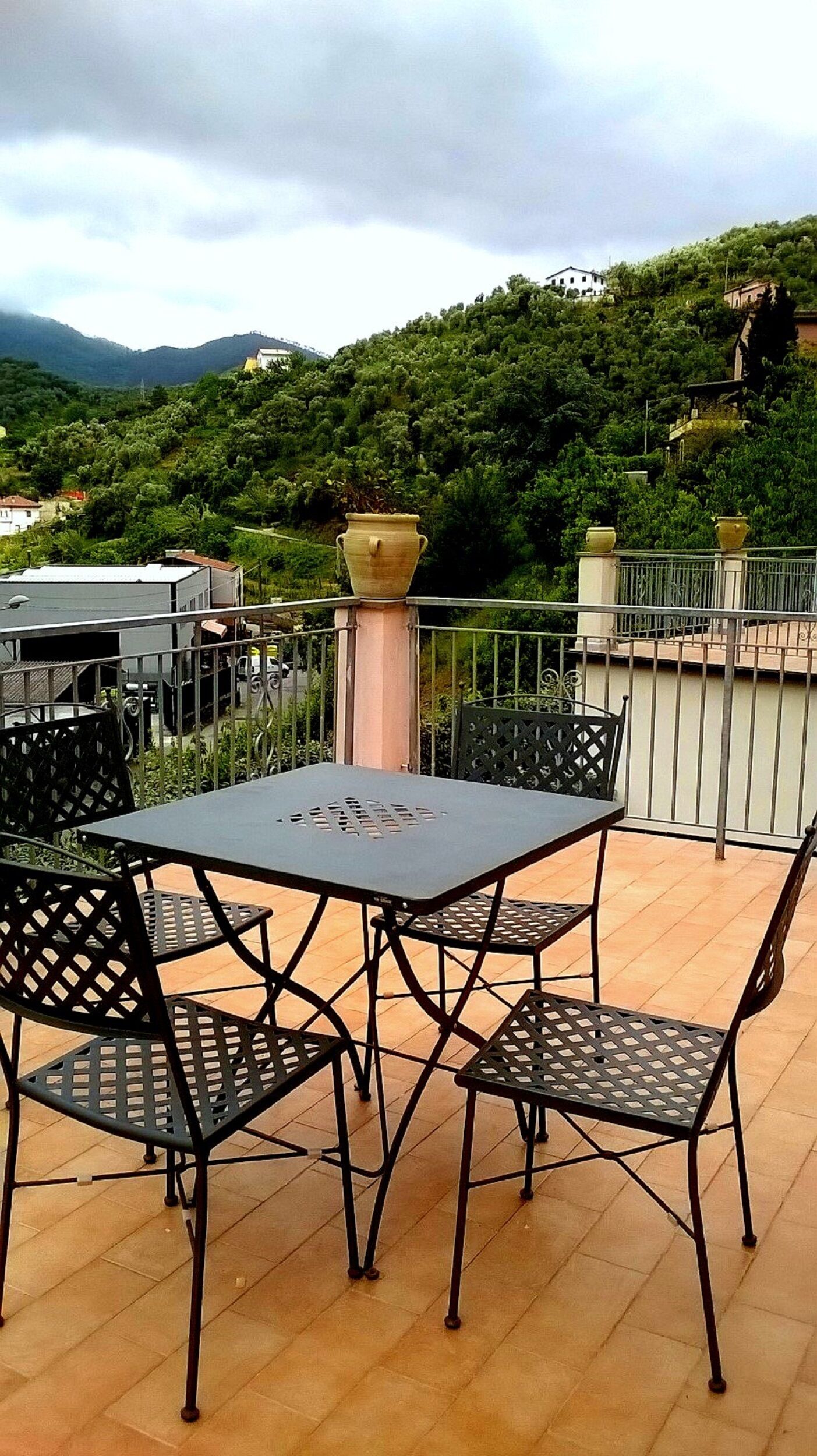 Um pátio externo com mesa e cadeiras, cercado por um guarda-corpo de metal, com vista para uma encosta coberta de vegetação e edifícios.