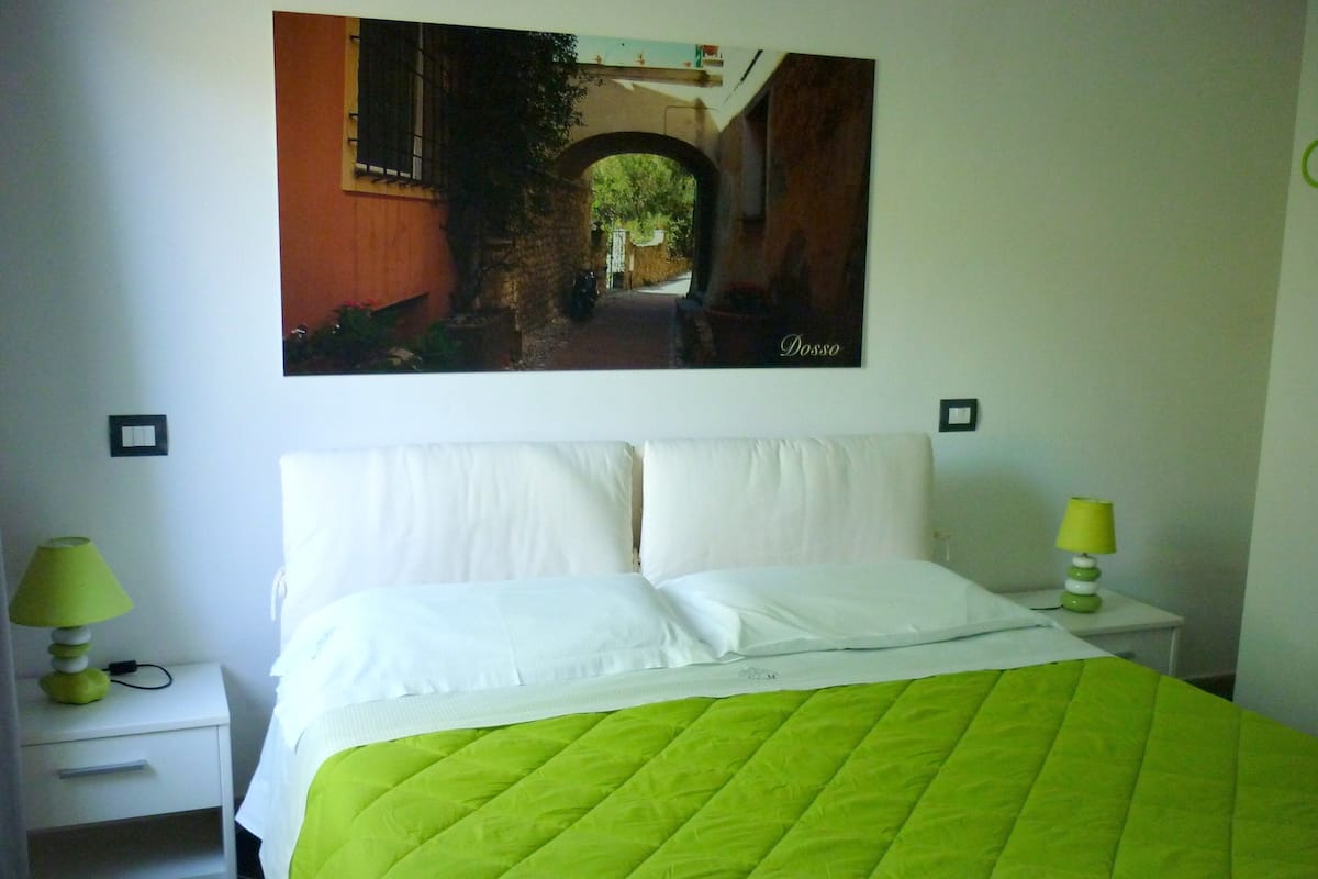 A bedroom with a bed, two bedside tables with lamps, a painting on the wall, and a tiled floor.