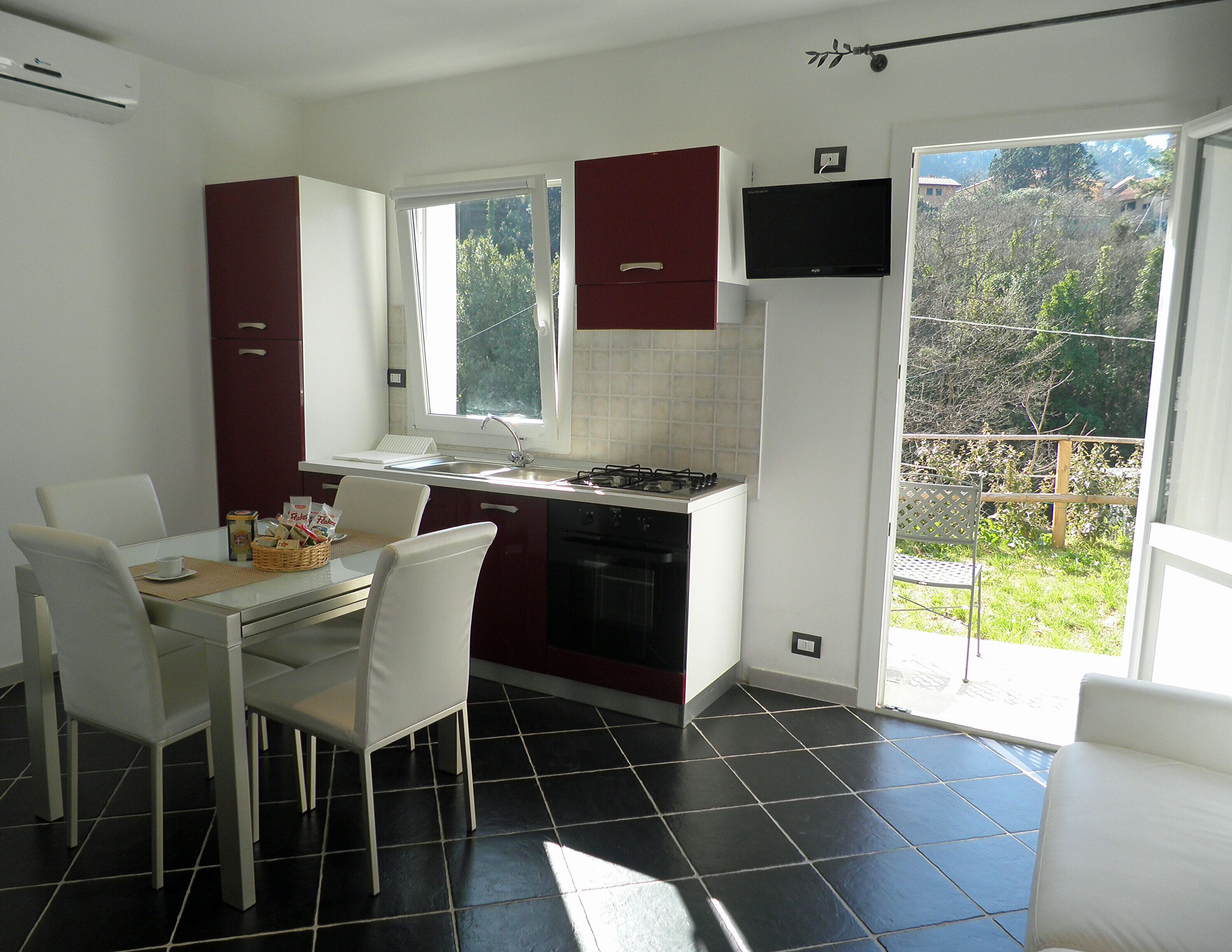 A compact kitchen with a dining area, a TV mounted above the stove, and a view of the outdoors through a sliding door.
