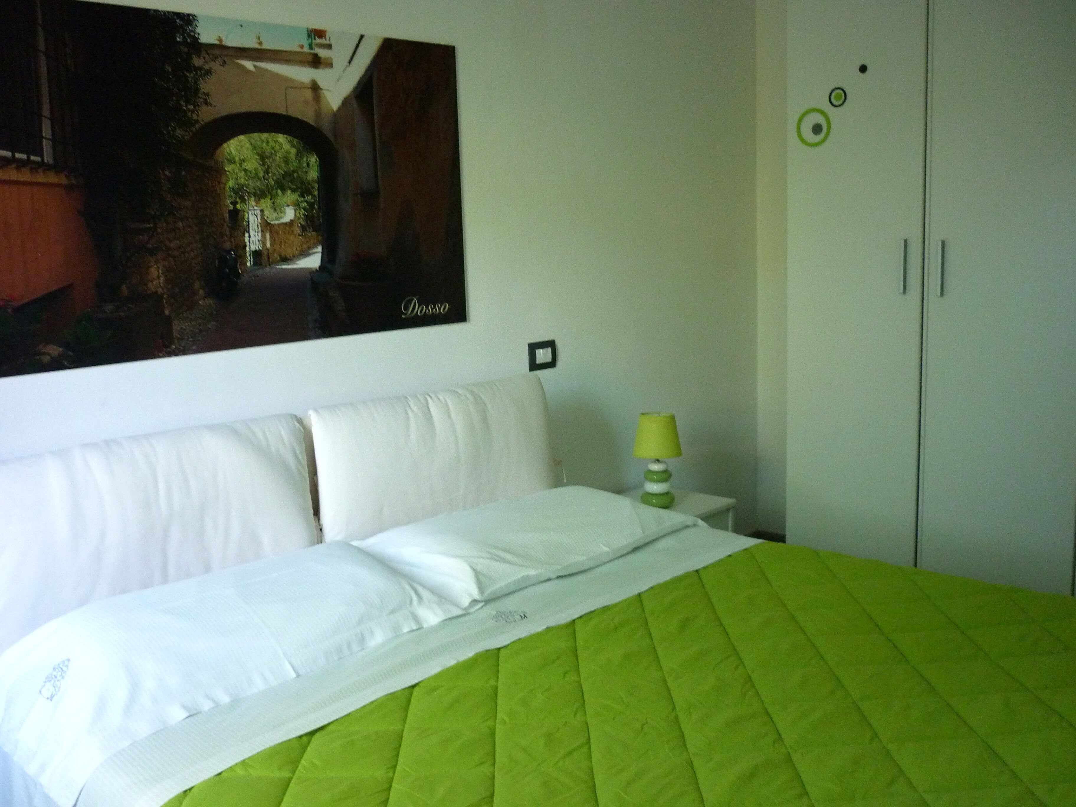 Una habitación de hotel con una cama, una colcha verde, almohadas blancas, una mesita de noche con una lámpara verde, un cuadro en la pared y un armario.