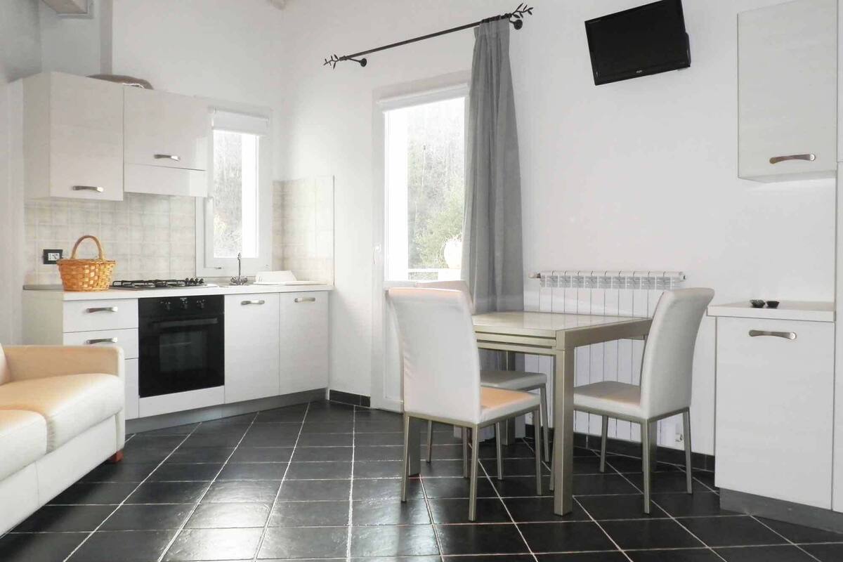 A modern kitchen with white cabinets, a dining table with chairs, a black tiled floor, and a window with a curtain.