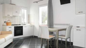 Eine moderne Küche mit weißen Schränken, einem Esstisch mit Stühlen, einem schwarzen Fliesenboden und einem Fenster mit Vorhang.
