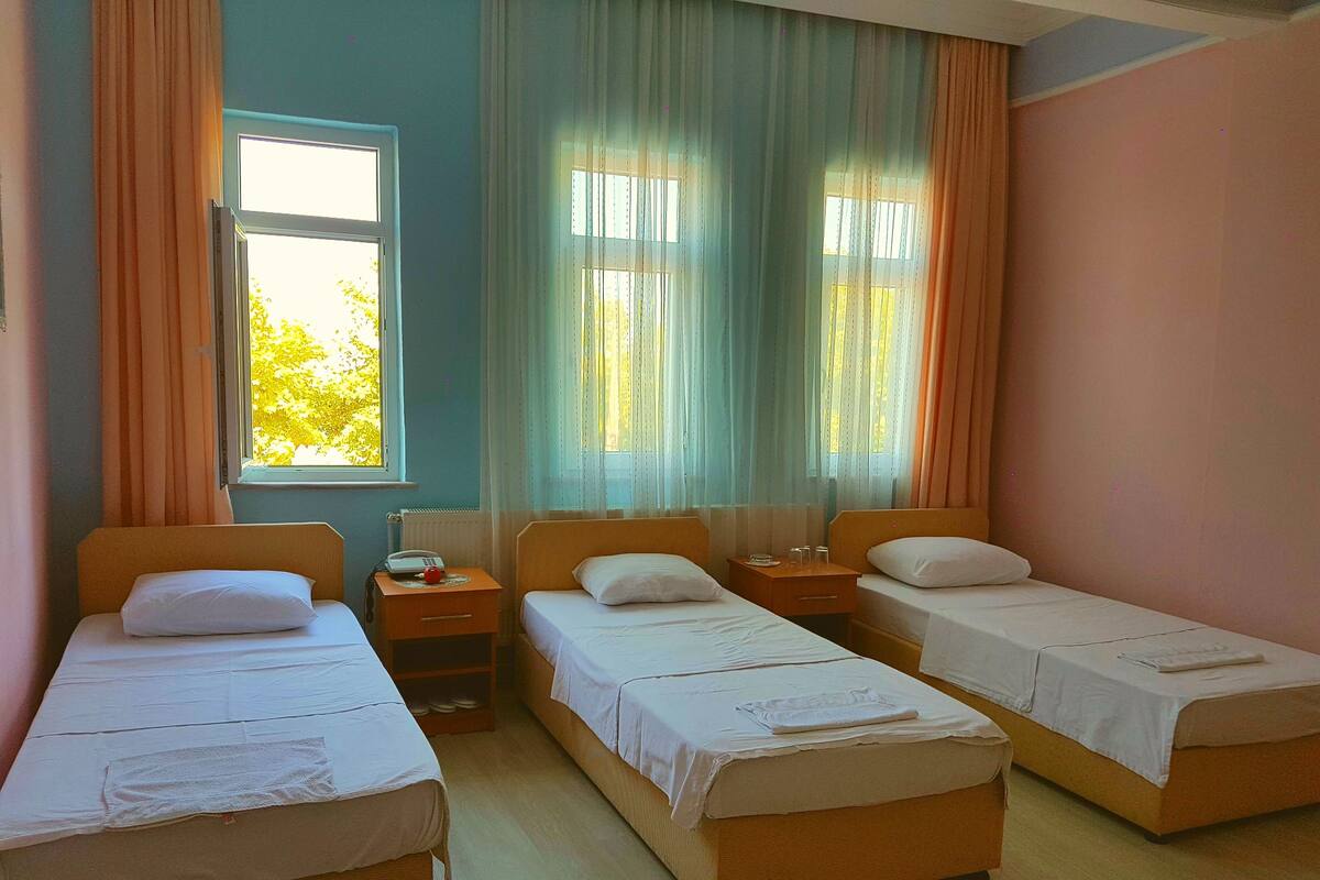 A room with three single beds, a window with curtains, and a bedside table with a phone and glasses.