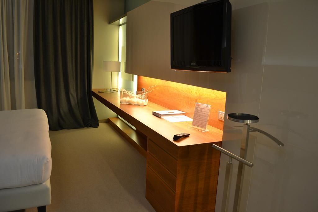 Quarto de hotel com escrivaninha de madeira, TV de tela plana acima dela, luminária e mesinha com bandeja.
