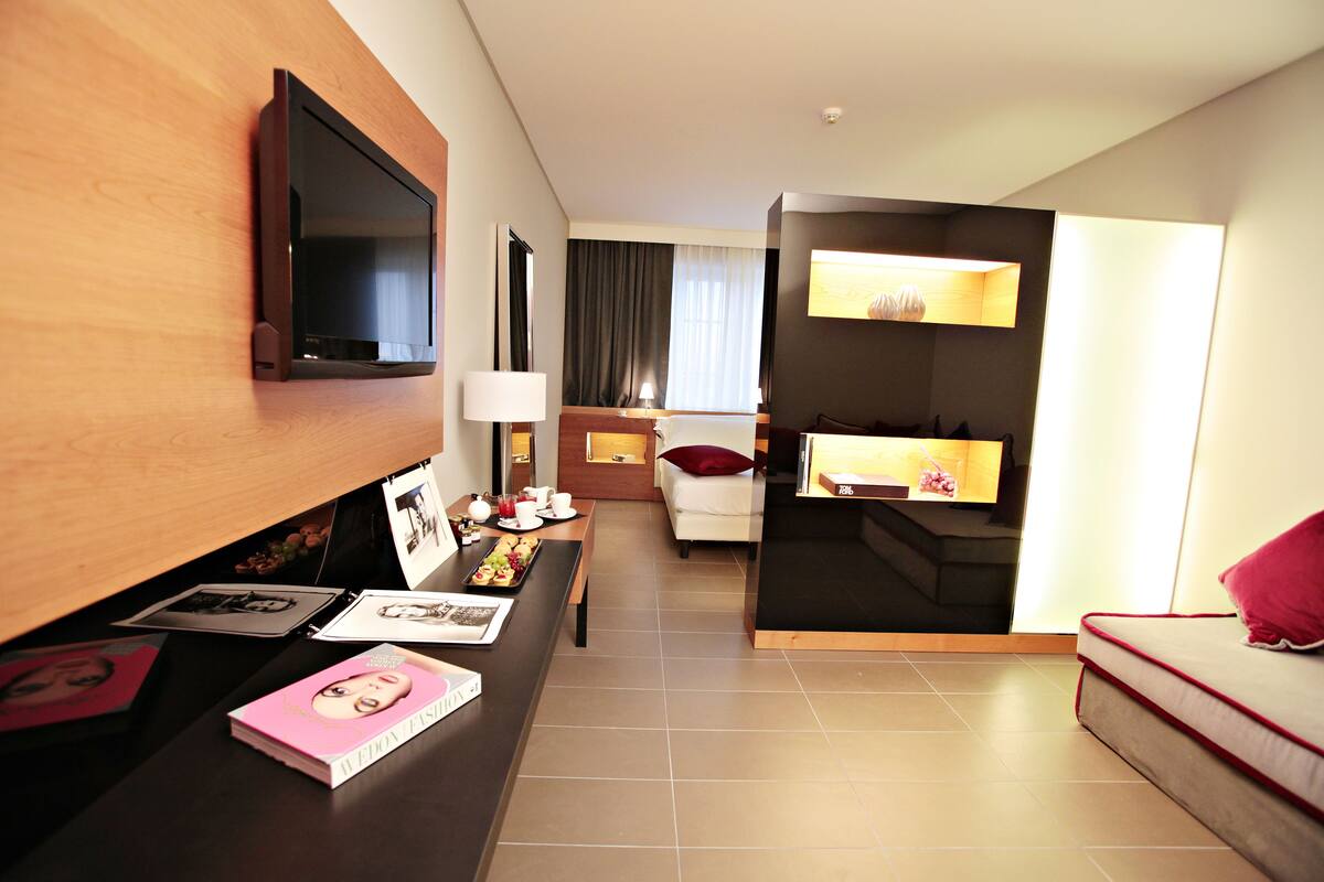 A hotel room with a flat-screen TV mounted on the wall, a desk with a lamp, a bed with a red pillow, and a small table with a tray of snacks.