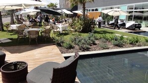 An outdoor pool area with lounge chairs and umbrellas, surrounded by a landscaped garden and a building with large windows. - Cosmopolitan Hotel (Civitanova Marche)