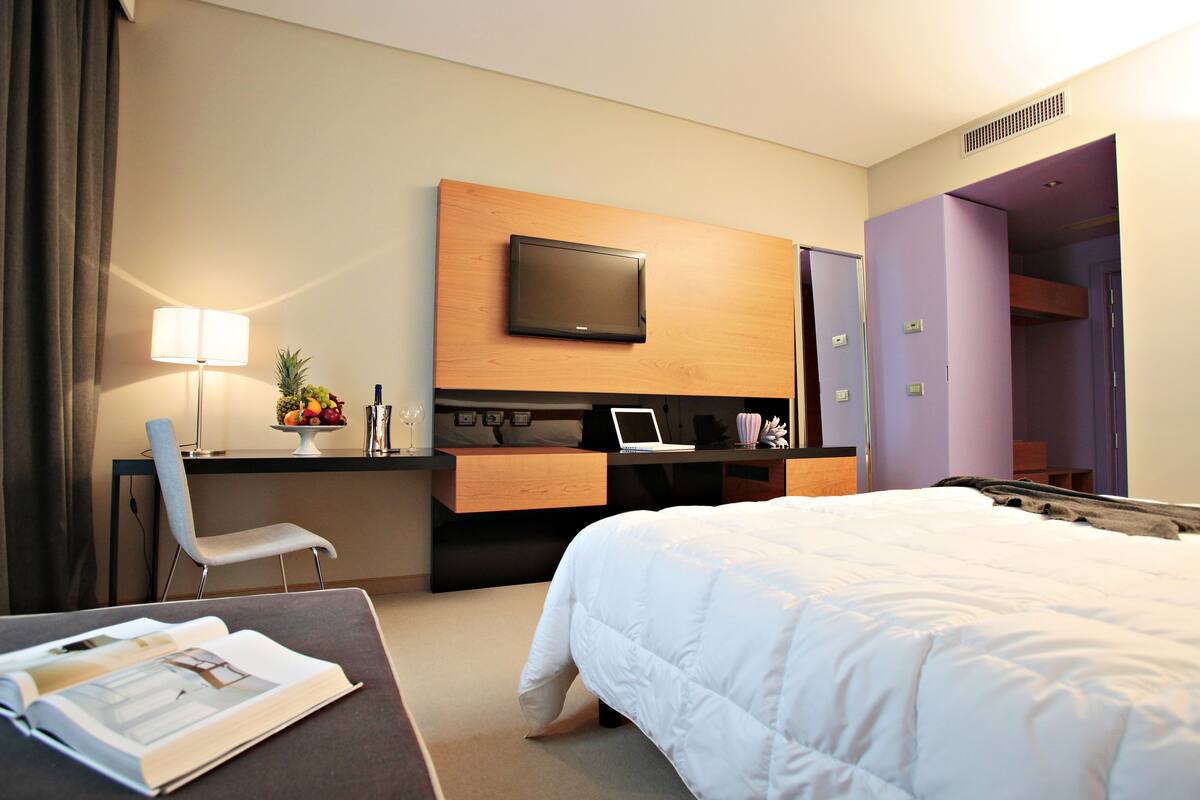 A hotel room with a bed, a desk with a laptop, a TV, a chair, and a lamp.
