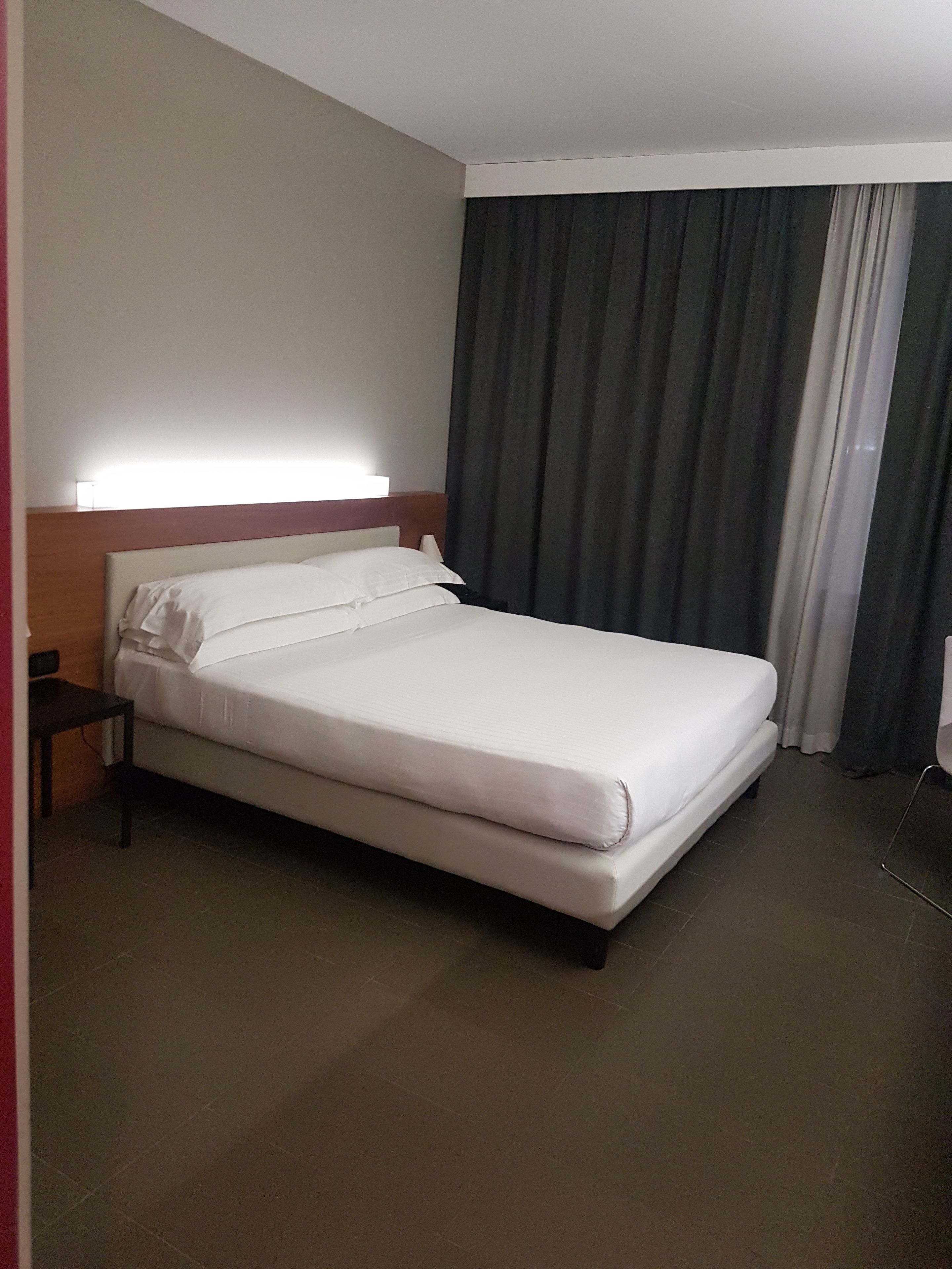 Une chambre d’hôtel avec un lit simple, une table de chevet, une lampe et une fenêtre avec des rideaux.