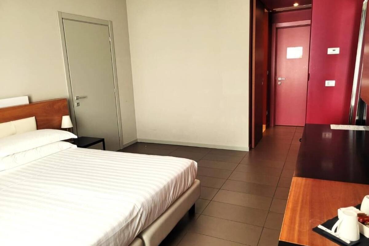 A hotel room with a bed, a nightstand, a wooden desk, and a red wall.