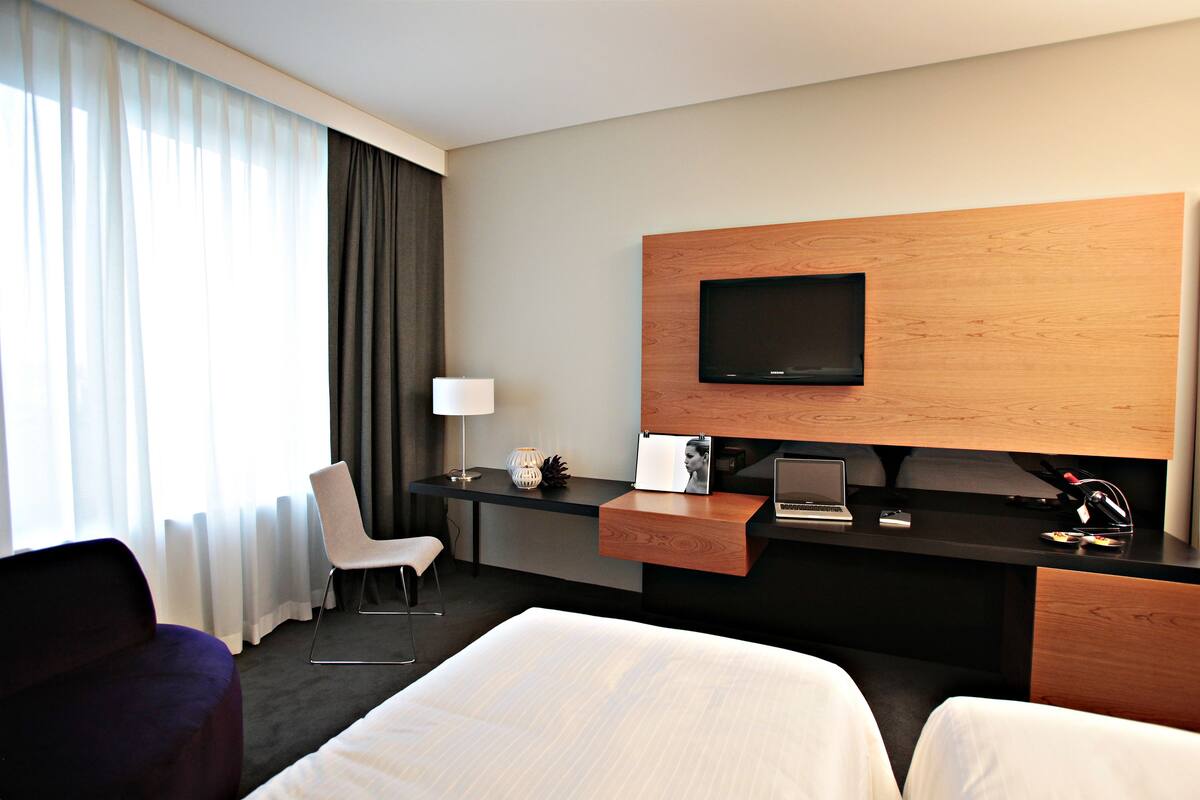 A modern hotel room with a large bed, a desk with a laptop, a television mounted on a wooden panel, and a chair.