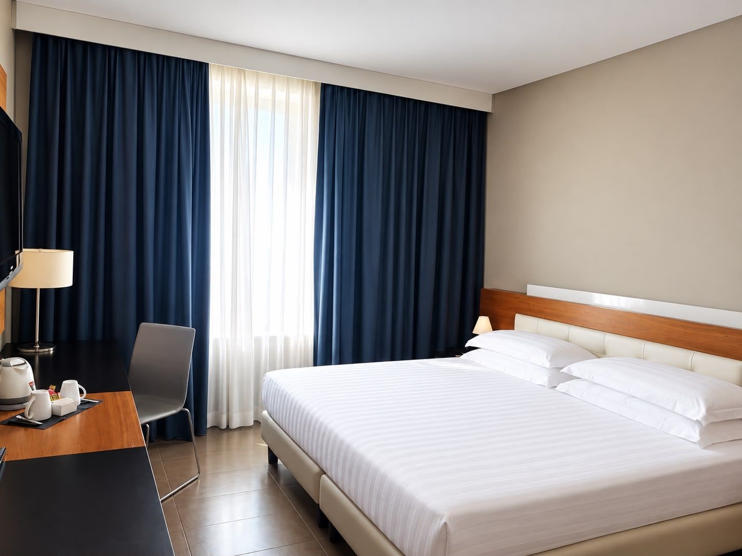 Habitación de hotel con cama, escritorio con lámpara, silla y ventana con cortinas.