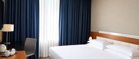 Habitación de hotel con cama, escritorio con lámpara, silla y ventana con cortinas.