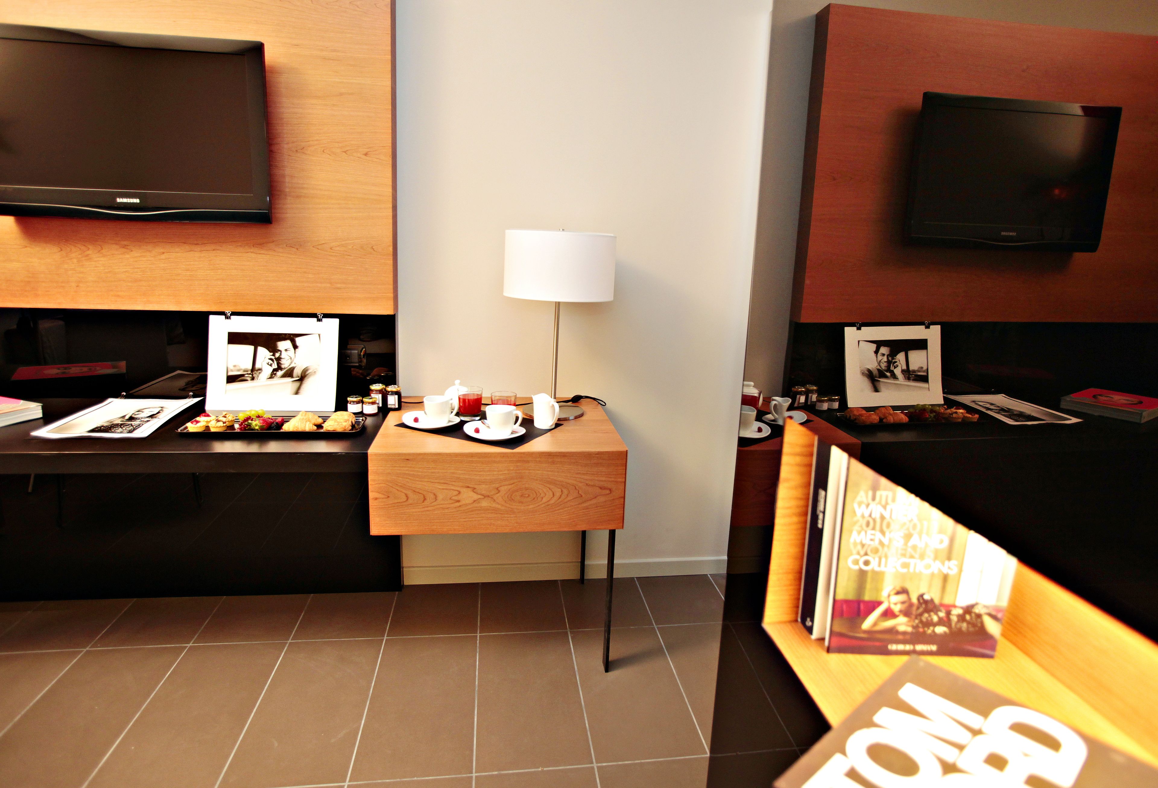 A hotel reception area with a wooden reception desk, a flat-screen TV mounted on the wall, a table with a lamp, and a display of books.