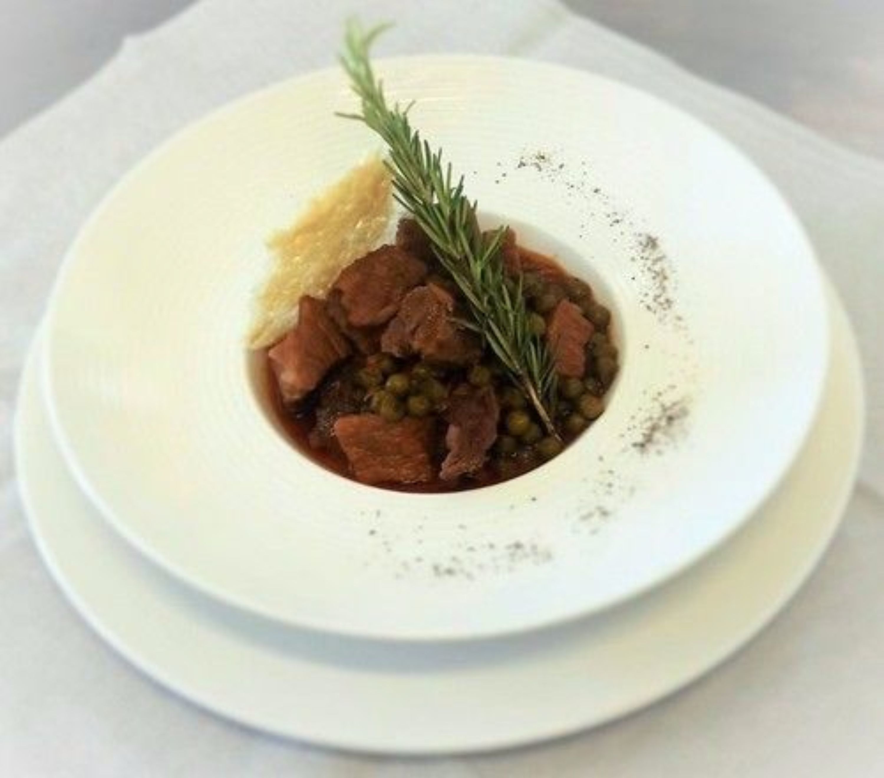 Prato branco com um prato de carne, ervilhas e um ramo de alecrim, servido sobre uma toalha de mesa branca.