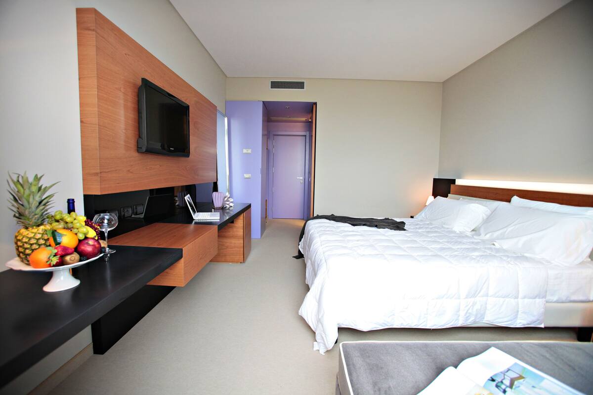 A hotel room with a bed, a desk with a computer, a TV mounted on the wall, and a fruit bowl on the desk.