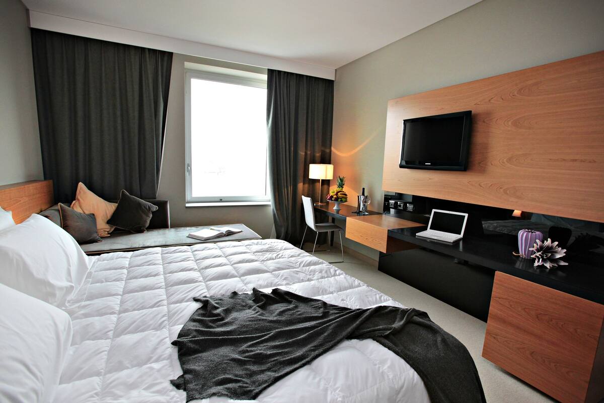 A hotel room with a bed, a desk with a laptop, a TV mounted on the wall, and a window with curtains.