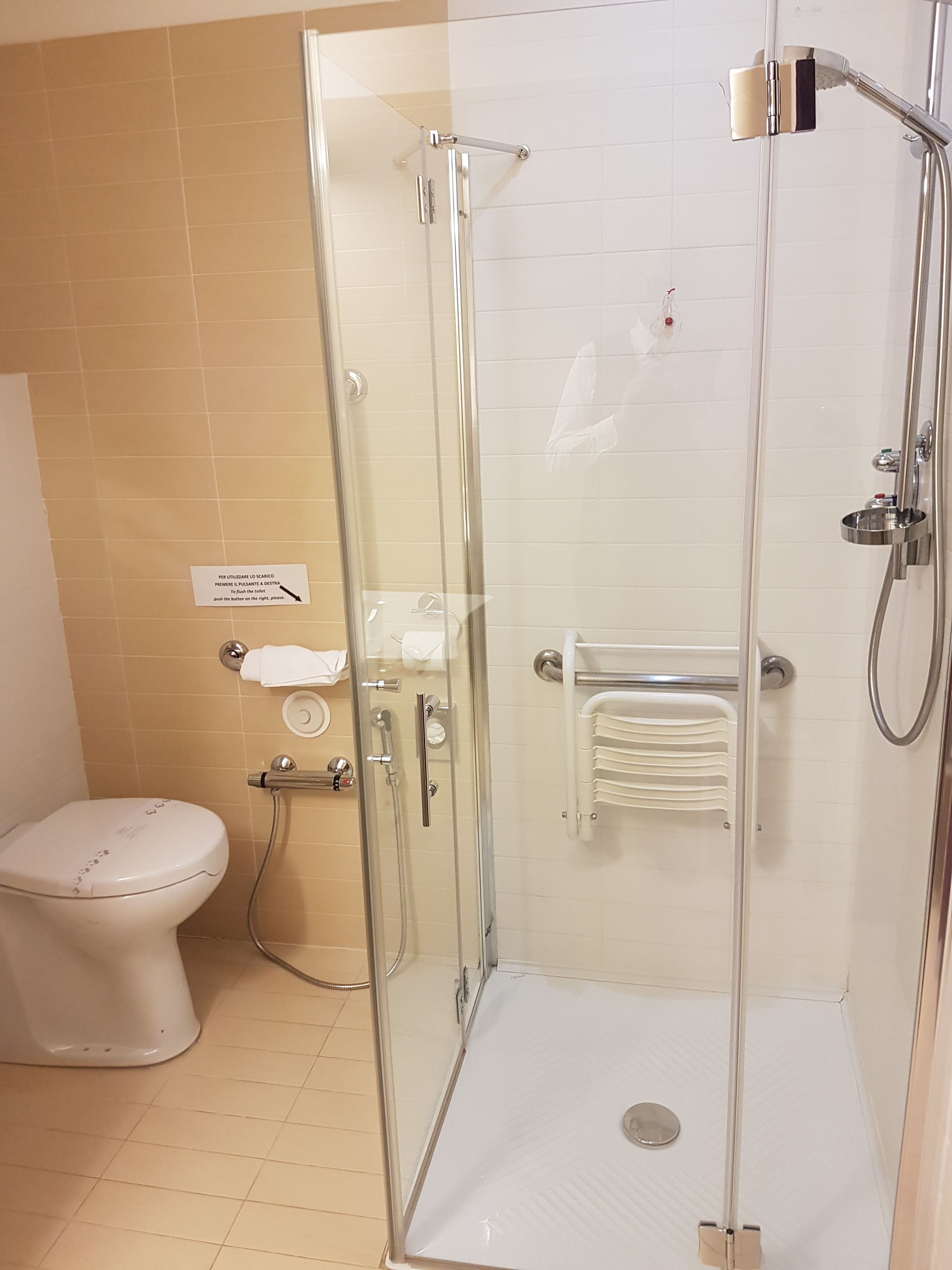 Cabina de ducha con paredes de vidrio, un cabezal de ducha de mano, un perchero y un inodoro con dispensador de toallas de papel.