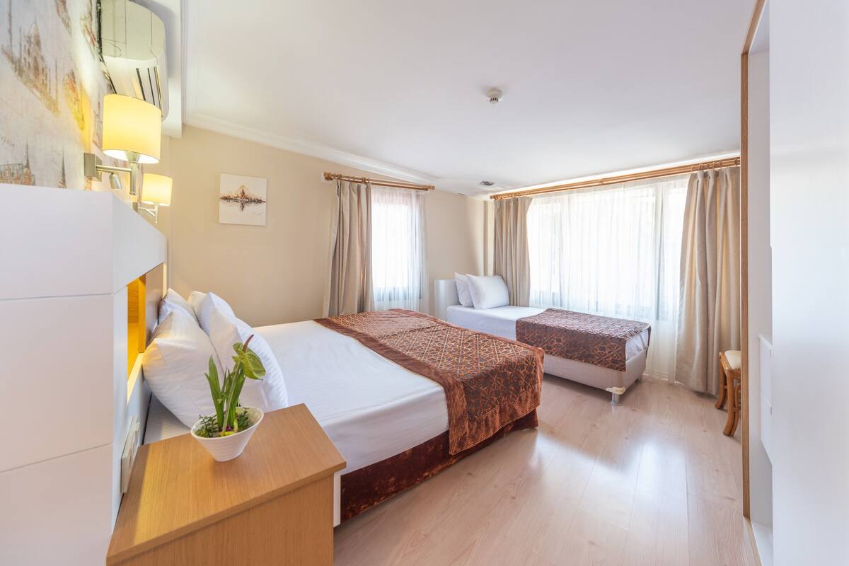 A hotel room with two beds, a bedside table with a plant, and a large window with curtains.