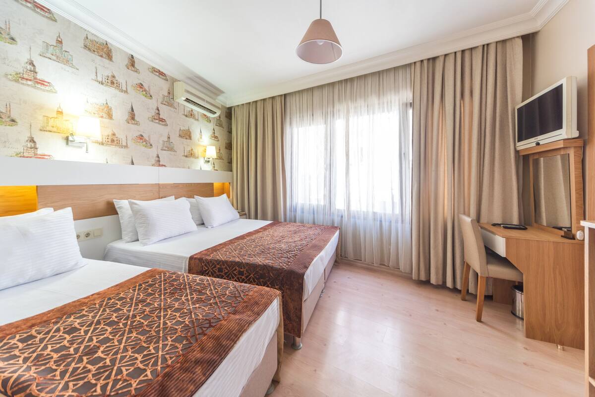 A hotel room with two beds, a desk, a chair, a TV, and a wall with cityscape wallpaper.