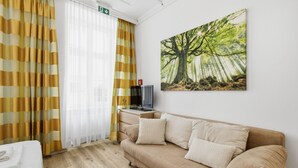 A room with a beige sofa, a television on a side table, and a painting of a tree on the wall. - AJO Vienna Messe - Contactless Check-in (Vienna)