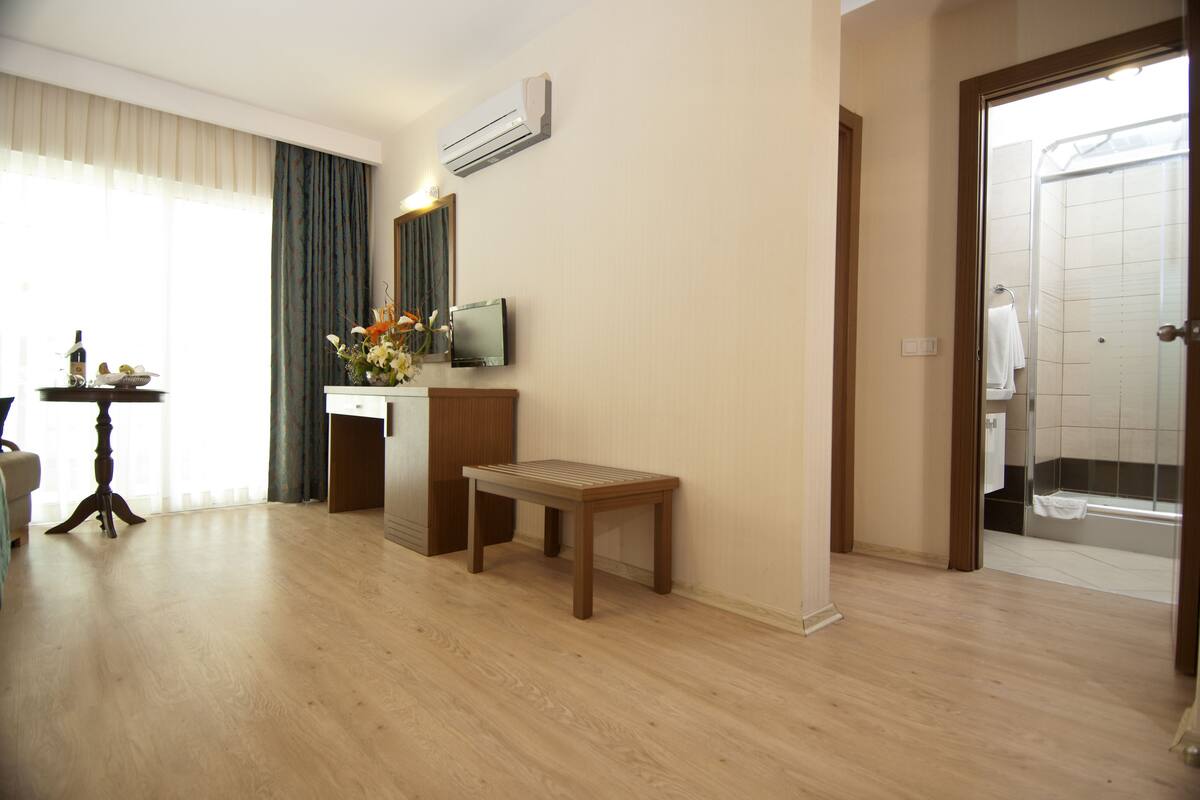 Une chambre d’hôtel avec un plancher en bois, un petit banc en bois, un téléviseur posé sur un meuble, un vase avec des fleurs et un espace douche visible à travers une porte ouverte.