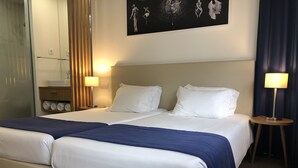 A double bed with white and blue bedding, two bedside lamps, a wooden nightstand, a wall-mounted shelf with towels, and a black and white artwork above the bed. - Porto Coliseum Hotel (Porto)