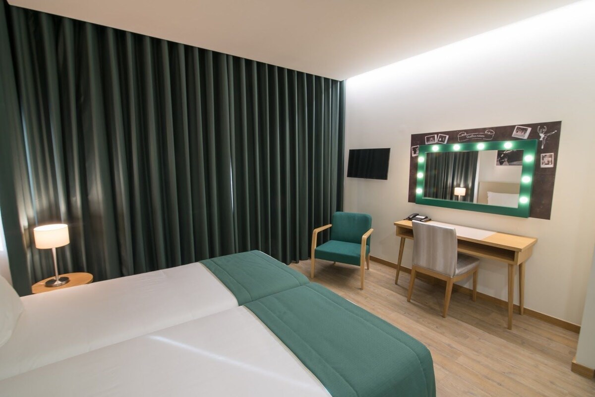 Habitación de hotel con cama, escritorio, silla, lámpara, televisión y espejo con luces.