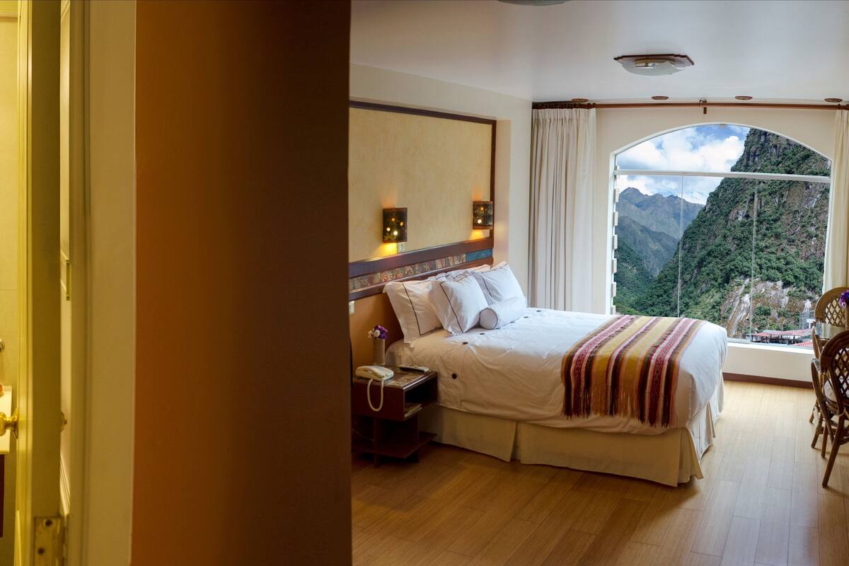 Un baño con un lavamanos de vidrio y un espejo, una mesita de noche con lámpara, y una ventana con vista a la montaña.