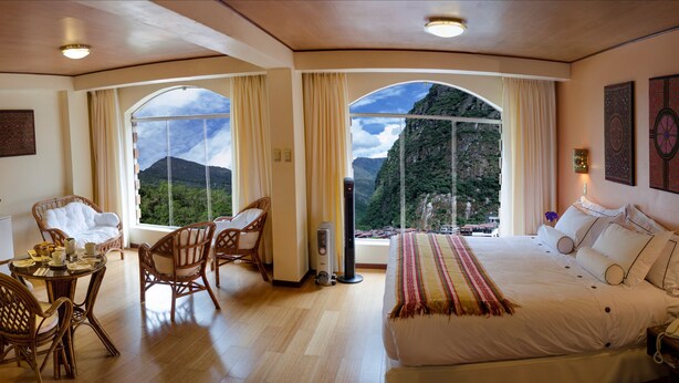 Ein Schlafzimmer mit einem großen Bett, einem Esstisch mit Stühlen und Blick auf Berge durch Rundbogenfenster.