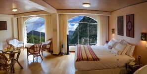 Une chambre avec un grand lit, une table à manger avec des chaises et une vue sur les montagnes, visible à travers des fenêtres en arc de cercle.
