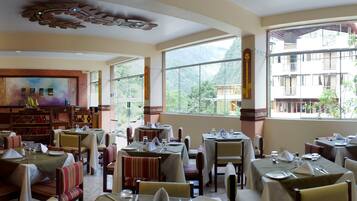 A restaurant with multiple tables and chairs, large windows with a mountain view, and a decorative ceiling.