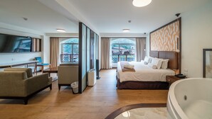 Ein Hotelzimmer mit einem großen Bett, einer Sitzecke mit Sofa und Sesseln, einem Fernseher, einer Badewanne und einem Balkon mit Blick.