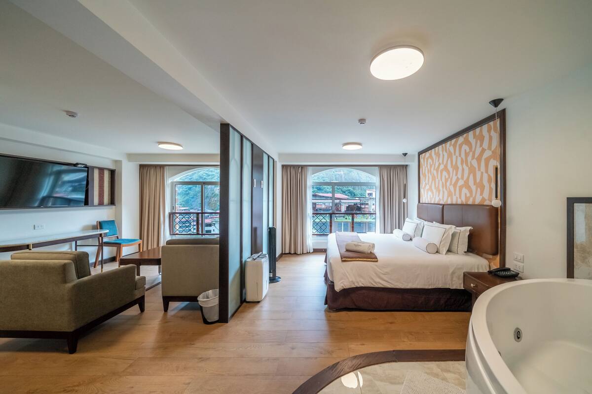 Habitación de hotel con una cama grande, una zona de estar con sofá y sillón, televisión, bañera y balcón con vistas.