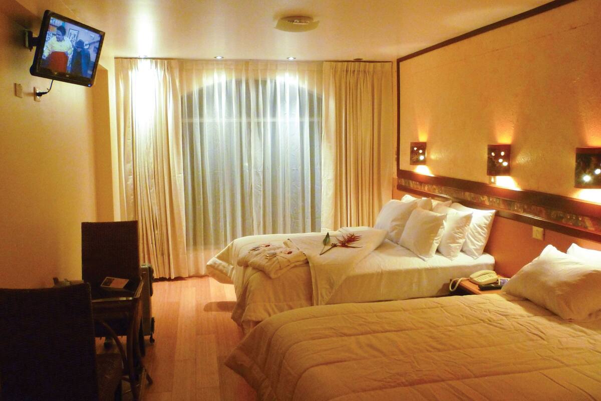 Habitación de hotel con dos camas, un televisor montado en la pared, un escritorio con una silla y una ventana con cortinas.