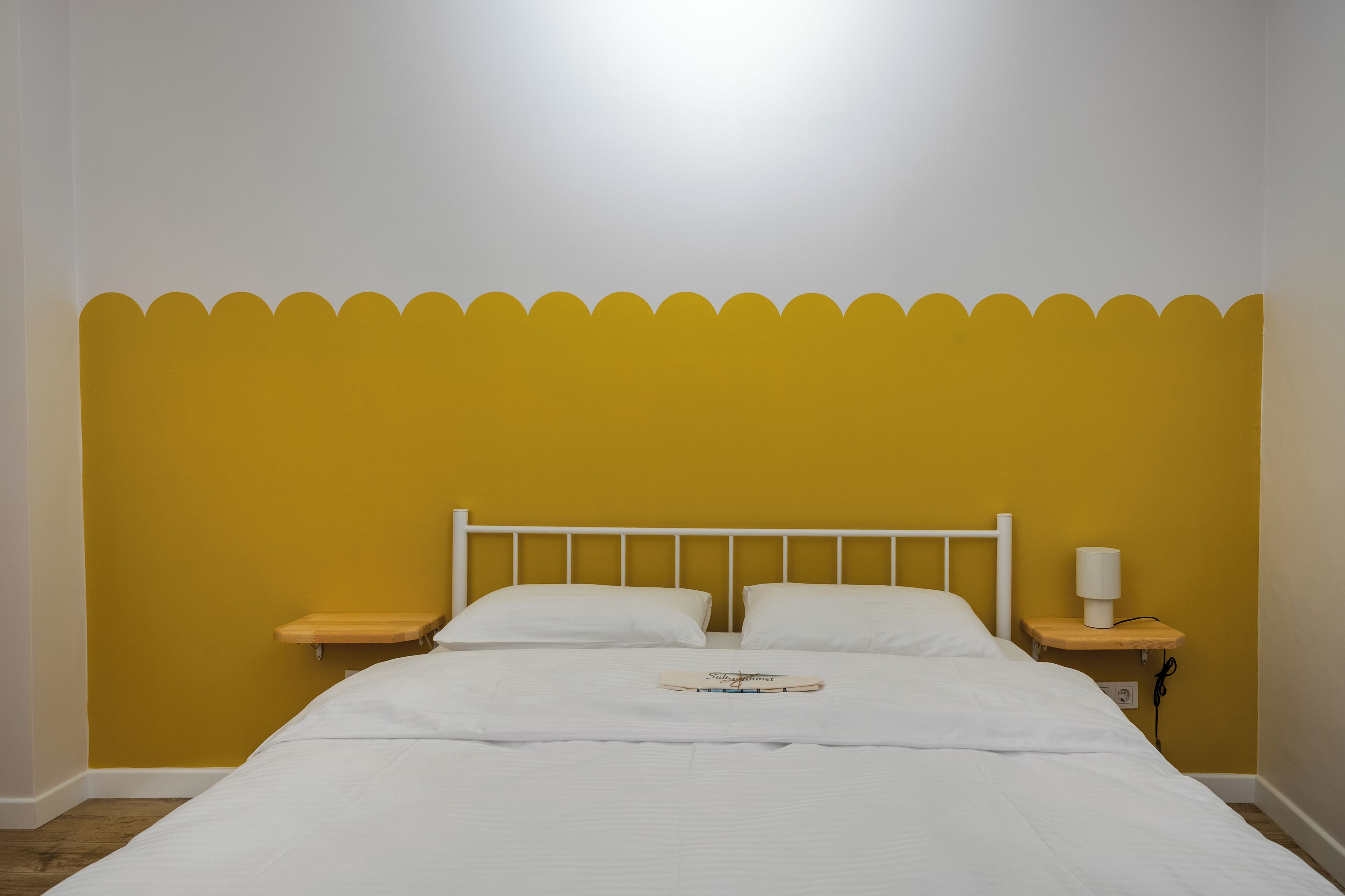 Una cama con ropa de cama blanca, un cabecero de madera, dos almohadas blancas, una mesita de noche con una lámpara y un pequeño estante.