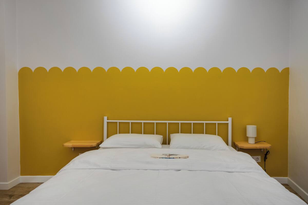 A bed with white bedding, a wooden headboard, two white pillows, a bedside table with a lamp, and a small shelf.