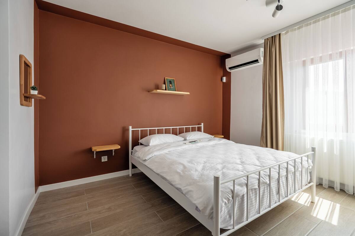 A bedroom with a white bed, brown walls, and a window with sheer curtains.