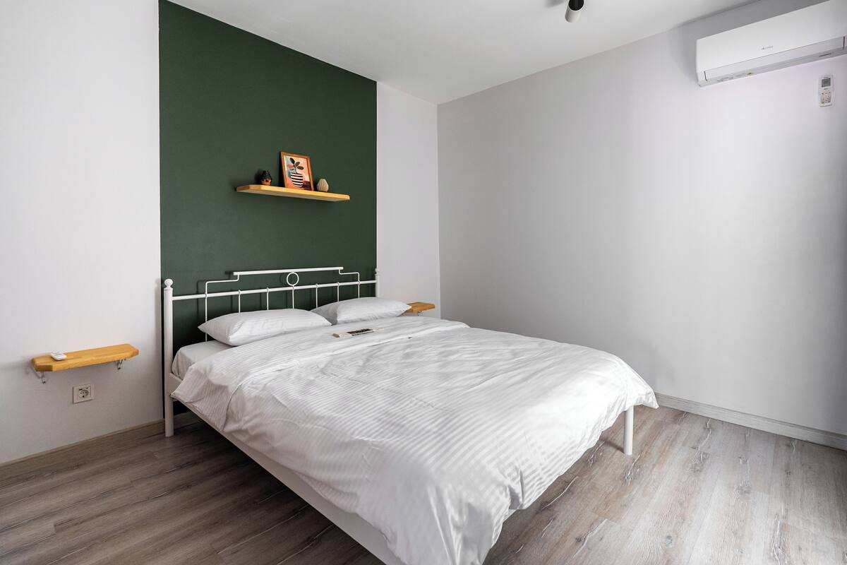 A bedroom with a bed, a green accent wall, a shelf with decorative items, a bedside table, and a wall-mounted air conditioner.