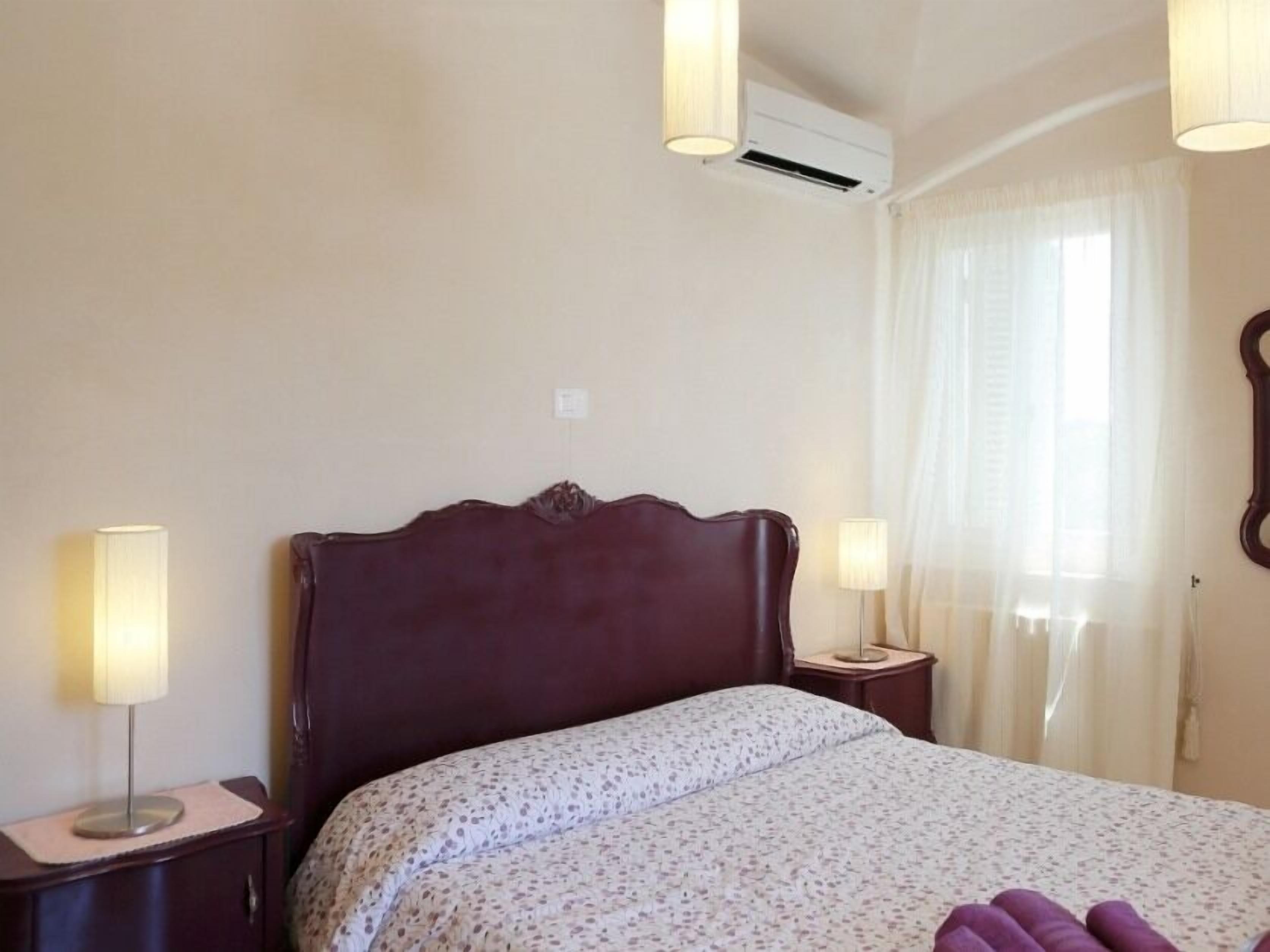 Habitación de hotel con cama, dos lámparas de noche, unidad de aire acondicionado, ventana con cortinas y espejo montado en la pared.